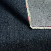 Close-up of dark indigo denim fabric with a folded corner revealing a lighter gray backside and a thin red and white edge stitch.