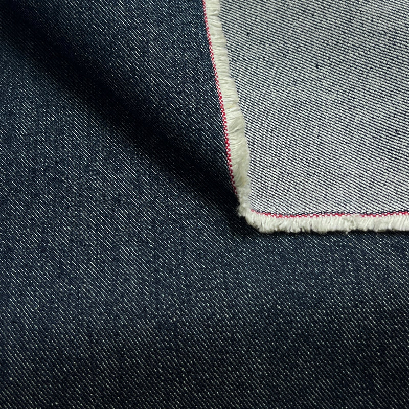 Close-up of dark indigo denim fabric with a folded corner revealing a lighter gray backside and a thin red and white edge stitch.