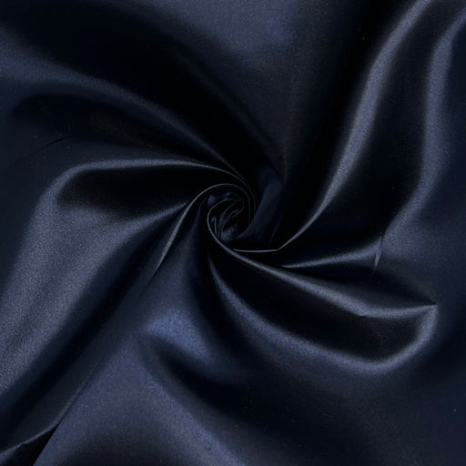 A close-up of dark navy blue satin fabric arranged in a spiral swirl, showing its smooth texture and subtle sheen.