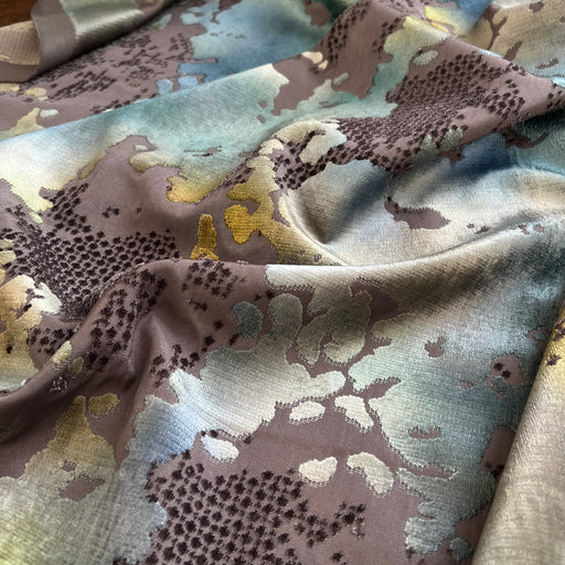 A close-up of draped fabric with an abstract floral or botanical pattern in muted taupe, dusty blue, pale green, and soft yellow, with darker brown dotted linework.