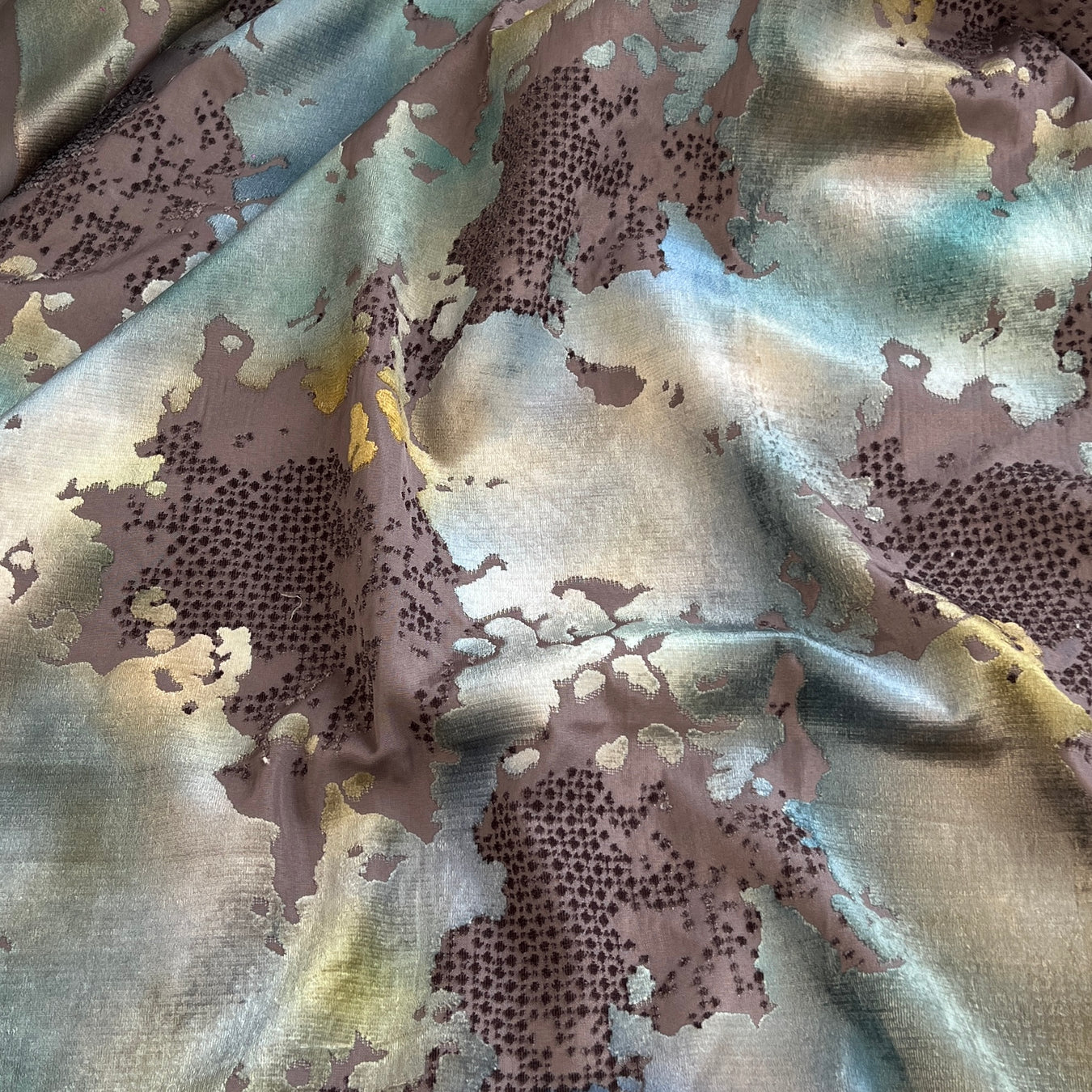 A close-up of draped fabric with an abstract, painterly pattern in muted taupe, pale blue, cream, and touches of yellow, with dark dotted texture in some areas.