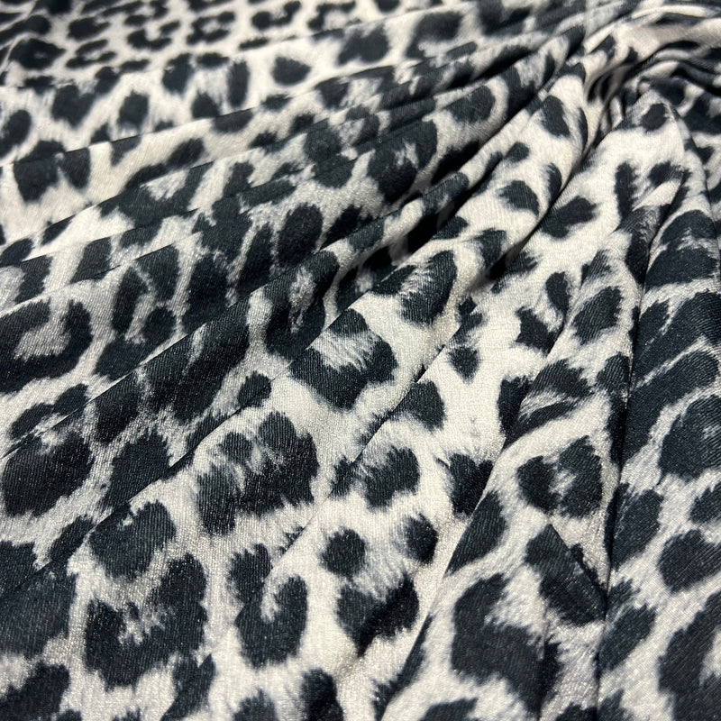 Leopard print fabric draped in soft folds, featuring a black and dark gray spotted pattern on a warm off-white background.