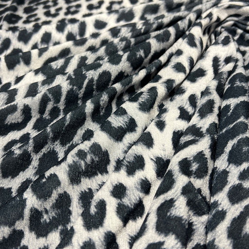 Leopard print fabric draped in soft folds, featuring a black and dark gray spotted pattern on a warm off-white background.