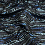 A close-up of a draped fabric with a dark charcoal base and horizontal stripes in metallic blue, light blue, and silver with geometric patterns.