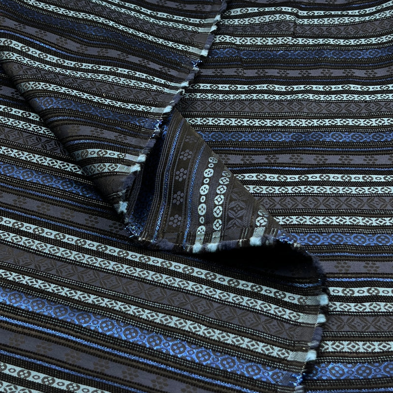 A close-up of patterned fabric with horizontal woven stripes in black, dark brown, blue, and off-white, shown with a folded corner revealing the texture.