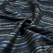 A close-up of draped fabric with horizontal wavy stripes in black, dark gray, metallic silver, and bright blue, featuring geometric and floral woven patterns.