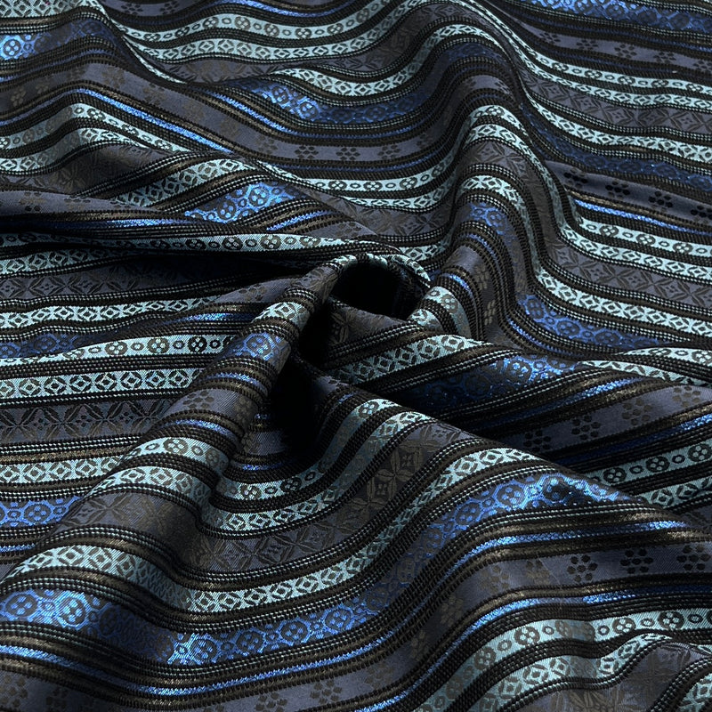 A close-up of draped fabric with horizontal wavy stripes in black, dark gray, metallic silver, and bright blue, featuring geometric and floral woven patterns.