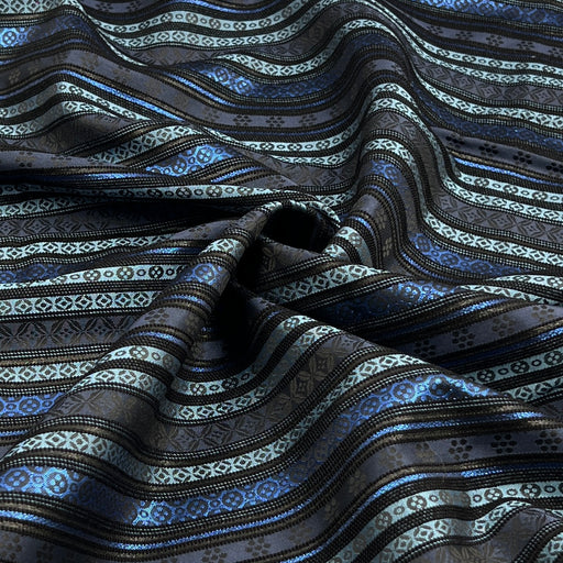 A close-up of draped fabric with horizontal wavy stripes in black, dark gray, metallic silver, and bright blue, featuring geometric and floral woven patterns.