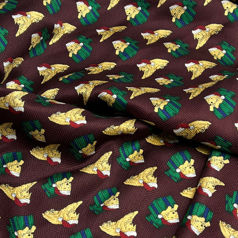 A close-up of draped fabric with a repeating holiday print featuring yellow puppies wearing red Santa hats and green plaid outfits on a dark brown background.