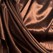 A close-up of draped satin fabric in a warm metallic bronze-brown color, showing the glossy sheen and soft folds.