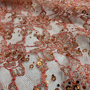 Close-up of a lace fabric featuring a white mesh base decorated with intricate coral-pink floral lace patterns, metallic bronze sequins, and small gold sequins.