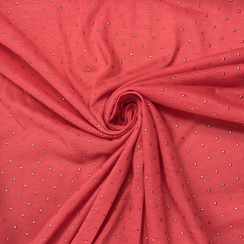 A close-up of coral pink fabric arranged in a spiral drape, featuring a repeating pattern of small metallic silver dots.