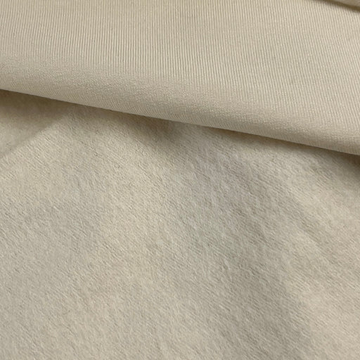 A close-up of a soft, textured fabric in a warm off-white or cream color, folded to show its ribbed and plush surface.
