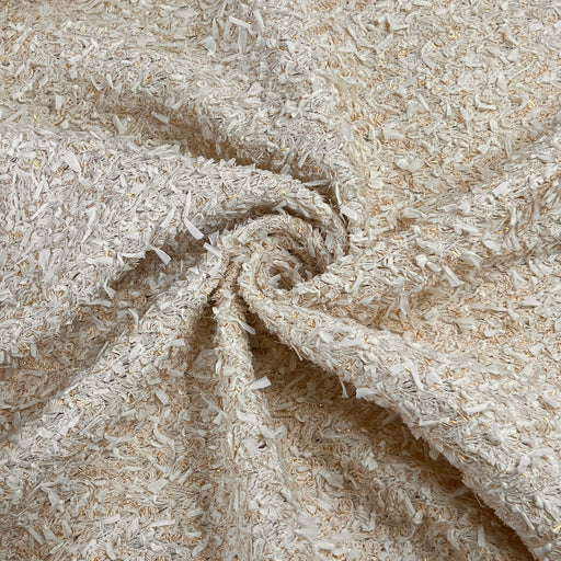 A close-up of a textured fabric with a shaggy, curly surface in warm off-white and beige tones, arranged in a spiral drape.