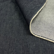 A close-up of dark indigo denim fabric with a folded corner revealing a lighter white underside and a finished raw edge.