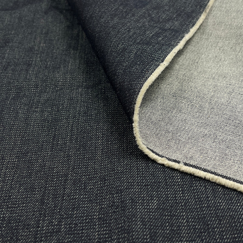 A close-up of dark indigo denim fabric with a folded corner revealing a lighter white underside and a finished raw edge.