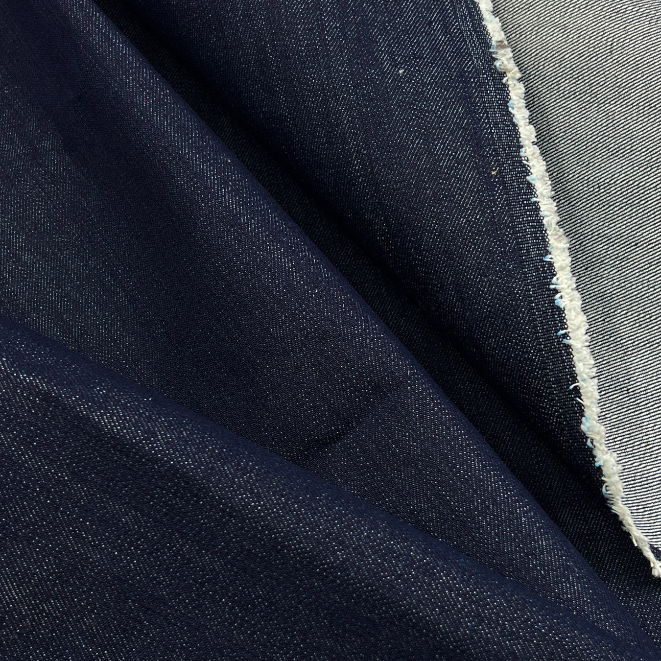 A close-up of dark indigo blue denim fabric with a visible white frayed edge showing the lighter inner weave.