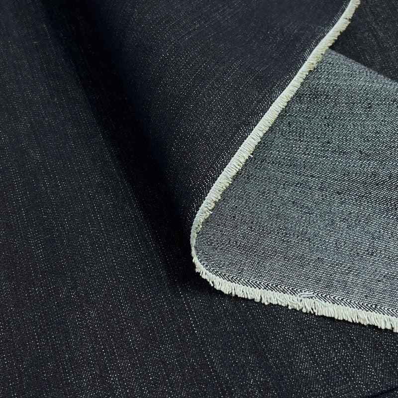 A close-up of dark indigo blue denim fabric with a folded corner revealing a lighter reverse side and a short selvedge.