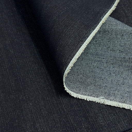A close-up of dark indigo blue denim fabric with a folded corner revealing a lighter reverse side and a short selvedge.