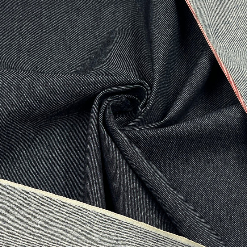 A close-up of dark indigo denim fabric with a diagonal twill texture, draped in a central swirl, with lighter gray fabric borders visible along the top right and bottom left edges.