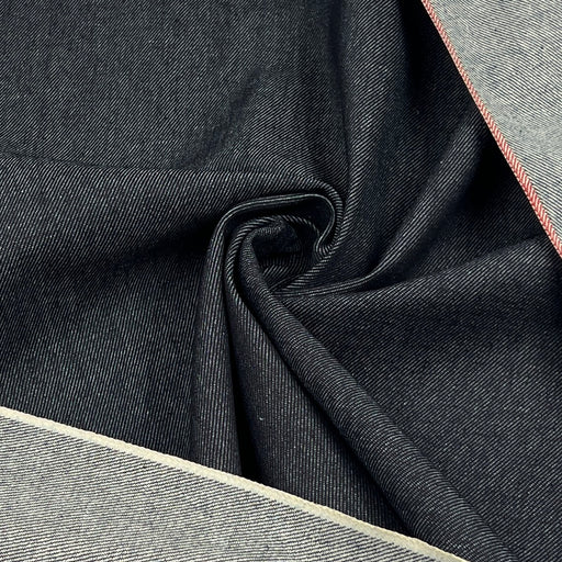 A close-up of dark indigo denim fabric with a diagonal twill texture, draped in a central swirl, with lighter gray fabric borders visible along the top right and bottom left edges.