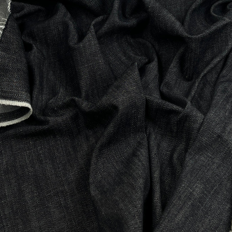 A close-up of dark indigo denim fabric with visible folds and a small portion of a light-colored hem or edge on the left side.
