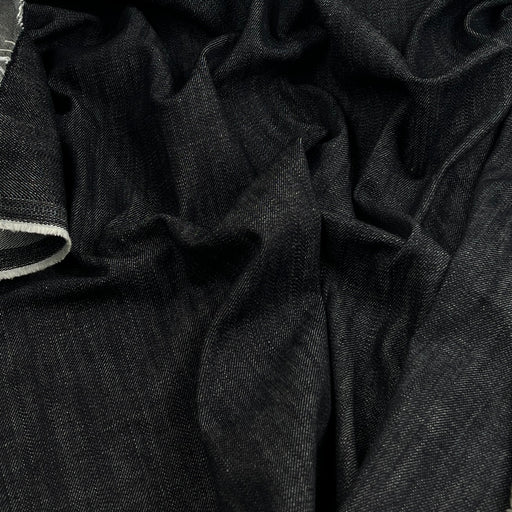 A close-up of dark indigo denim fabric with visible folds and a small portion of a light-colored hem or edge on the left side.