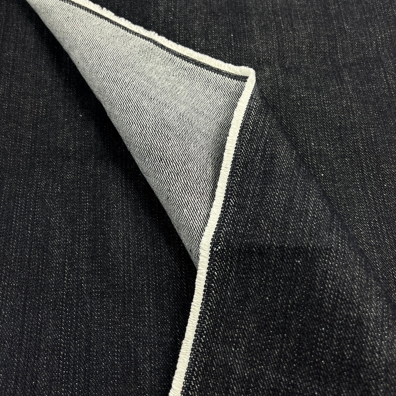 A close-up of dark indigo denim fabric with a folded corner revealing a light gray underside and a white stitched edge.
