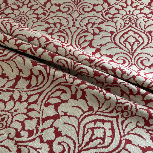 A close-up of patterned fabric with an ornate damask-style floral motif in deep red on a warm off-white or cream background, shown with soft folds.