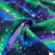 A close-up of a soft fleece fabric with a galaxy print, featuring swirling patterns of green, purple, blue, and black with white star details, draped to show its texture and color transitions.