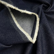 A close-up of dark indigo denim fabric with a white frayed selvedge edge and a lighter gray inner lining.