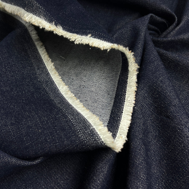 A close-up of dark indigo denim fabric with a white frayed selvedge edge and a lighter gray inner lining.