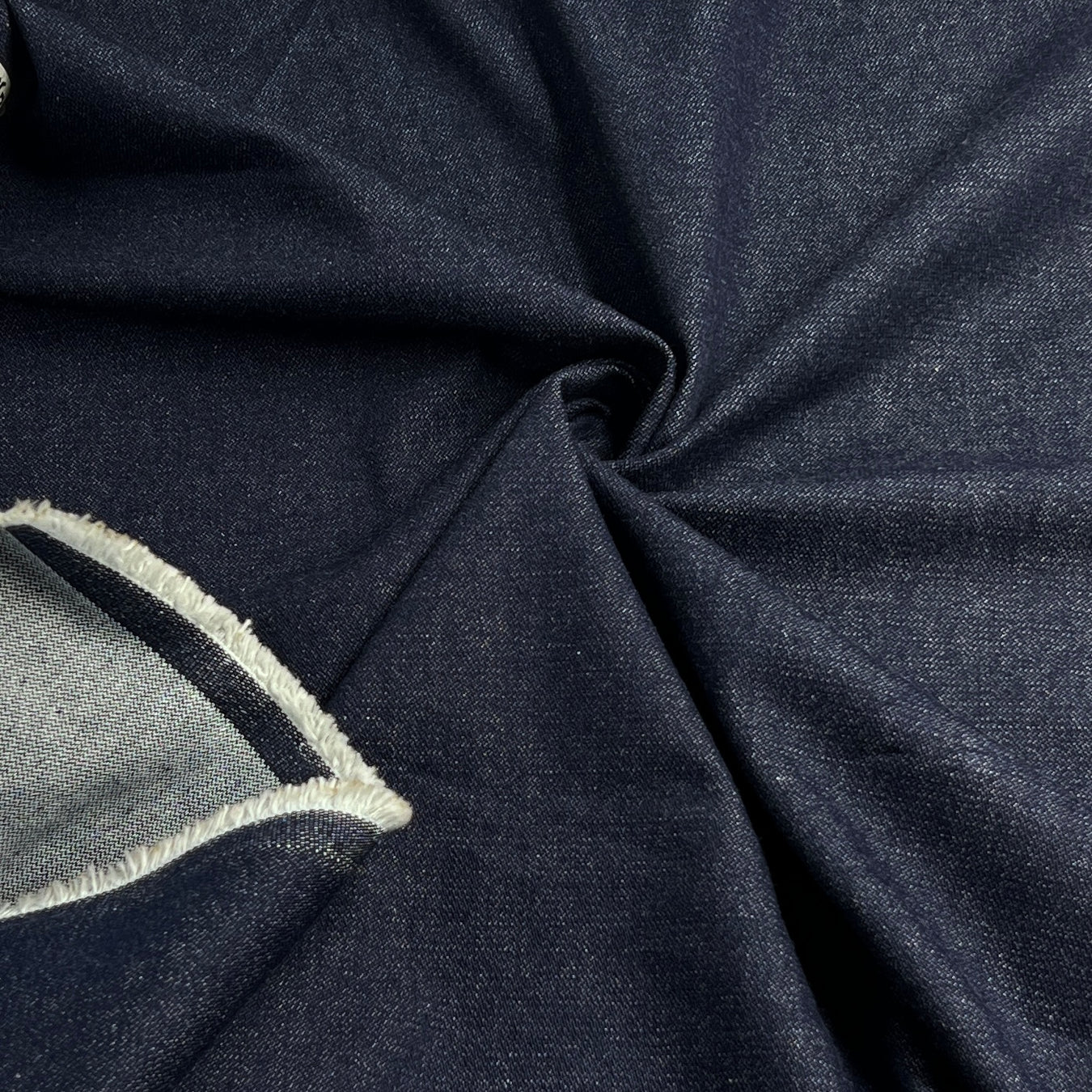 A close-up of dark navy blue fabric with a textured weave, draped in soft folds. A folded section on the left reveals a lighter gray inner layer with a dark navy stitched edge.