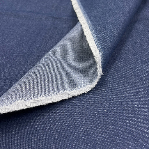 A close-up of dark indigo denim fabric with a folded corner showing the lighter blue reverse side and frayed white edges.