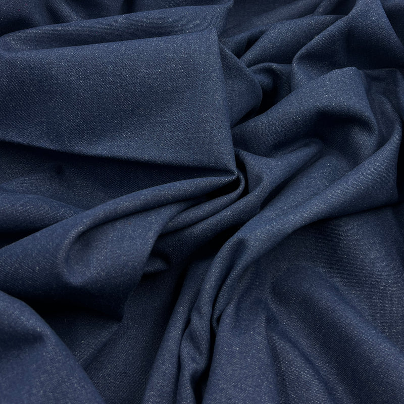 A close-up of draped indigo blue denim fabric with a fine textured weave, showing soft folds and shadows.