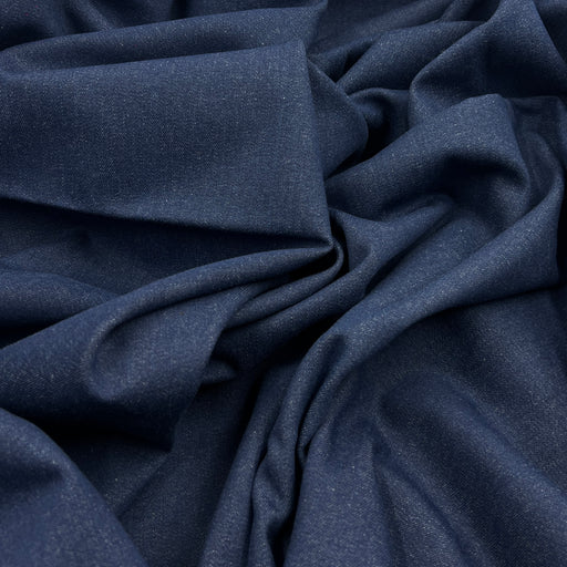 A close-up of draped indigo blue denim fabric with a fine textured weave, showing soft folds and shadows.