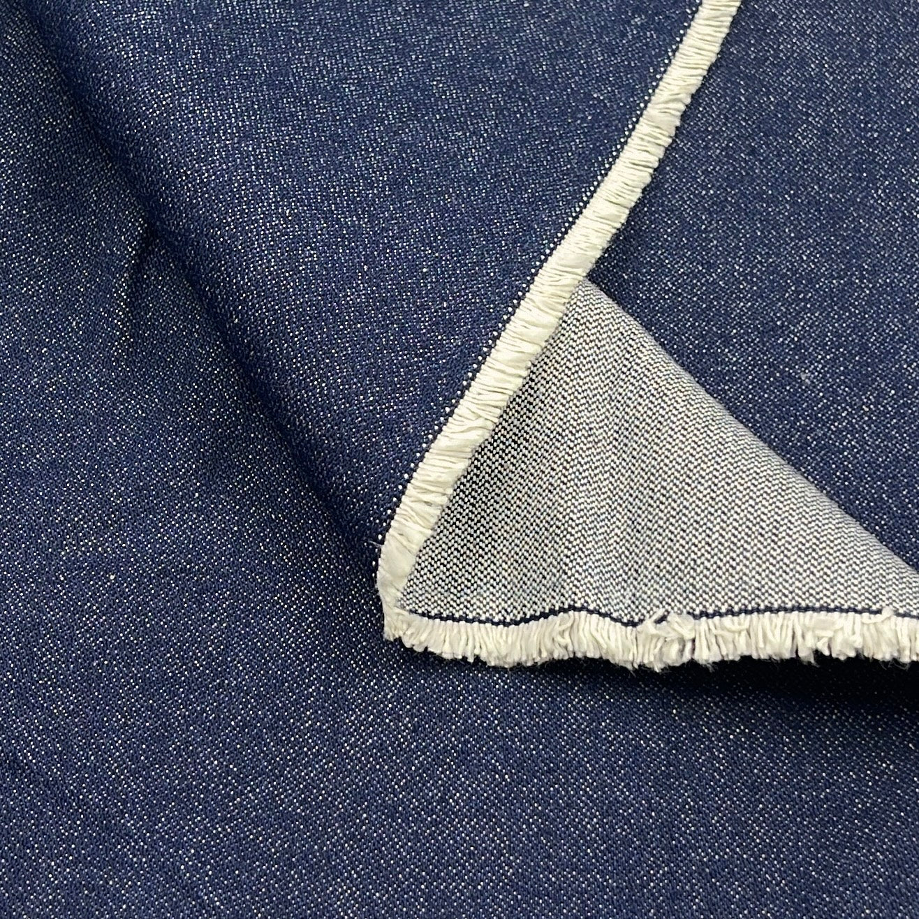 A close-up of dark blue denim fabric with a folded corner showing a light beige frayed edge.