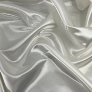 A close-up of draped satin fabric with a soft sheen, showing folds and a light warm gray to off-white color.