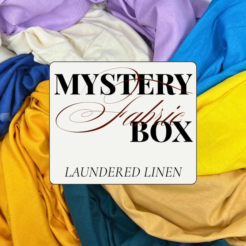 A white rectangular label reading 'MYSTERY Fabric BOX' and 'LAUNDERED LINEN' placed over a pile of colorful fabric swatches in purple, blue, cream, yellow, and teal.