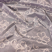 A close-up of a sheer, shimmering lavender-gray fabric with a repeating white outline pattern of flowers and swirls.