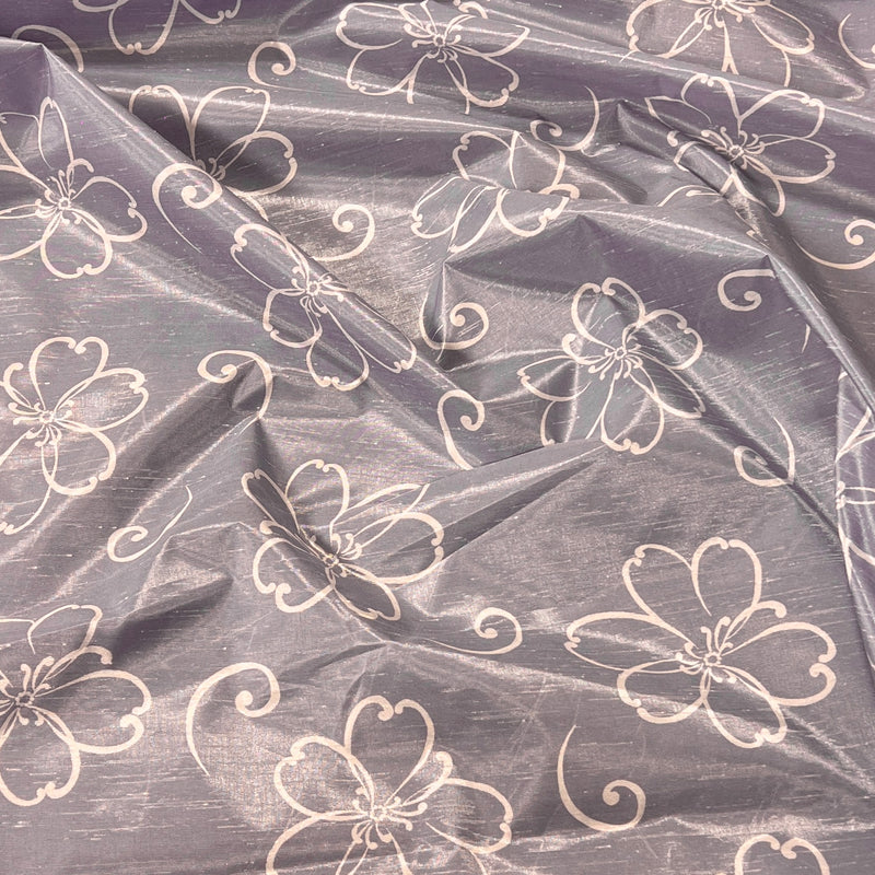 A close-up of a sheer, shimmering lavender-gray fabric with a repeating white outline pattern of flowers and swirls.