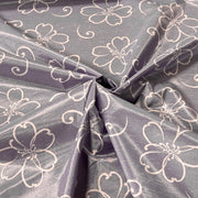 A close-up of draped fabric with a lavender-gray base and a repeating white floral line pattern, gathered in the center to show the material's sheen and drape.