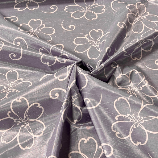 A close-up of draped fabric with a lavender-gray base and a repeating white floral line pattern, gathered in the center to show the material's sheen and drape.