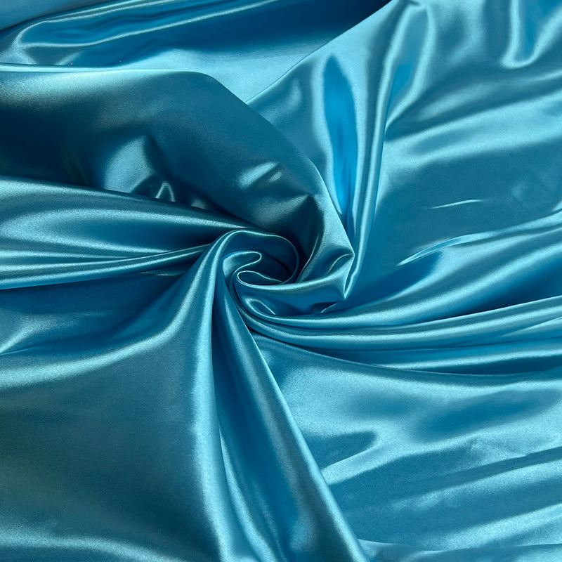 A close-up of shiny blue satin fabric arranged in soft folds and swirls, showing the material's lustrous texture and sheen.