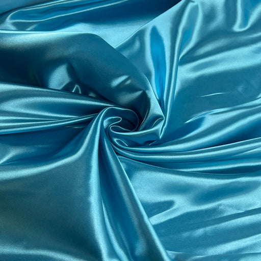 A close-up of shiny blue satin fabric arranged in soft folds and swirls, showing the material's lustrous texture and sheen.
