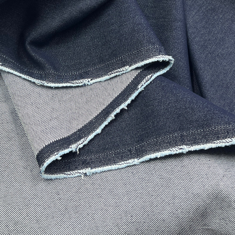 A close-up of folded dark indigo denim fabric with a raw, frayed selvedge edge showing the lighter inner weave.