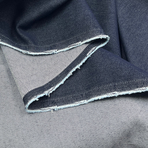 A close-up of folded dark indigo denim fabric with a raw, frayed selvedge edge showing the lighter inner weave.