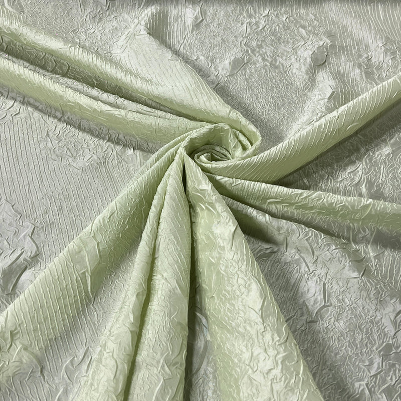 A close-up of pale sage green textured taffeta fabric with a textured floral pattern, draped and swirled to show its texture and sheen.