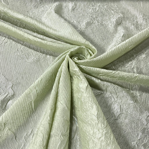 A close-up of pale sage green textured taffeta fabric with a textured floral pattern, draped and swirled to show its texture and sheen.