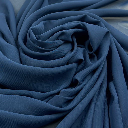 Closeup of sheer polyester chiffon fabric in navy blue pulled into a swirl showing off fluid drape. Photographed over white background for contrast. 